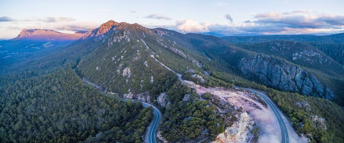 A winding mountain road through a dense forest with a prominent peak in the background.