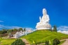 A large white statue of a seated figure on a hill, surrounded by greenery and a clear blue sky.