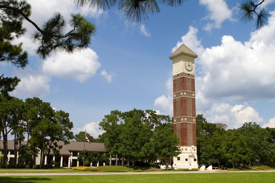 A clock tower with a clock face, surrounded by trees and a building with a flag.