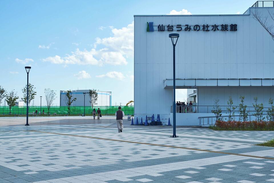 A modern building with a sign in Japanese, a tiled plaza, and a few people walking.