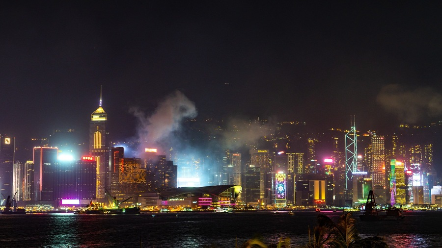 A nighttime cityscape with illuminated skyscrapers and a body of water in the foreground.