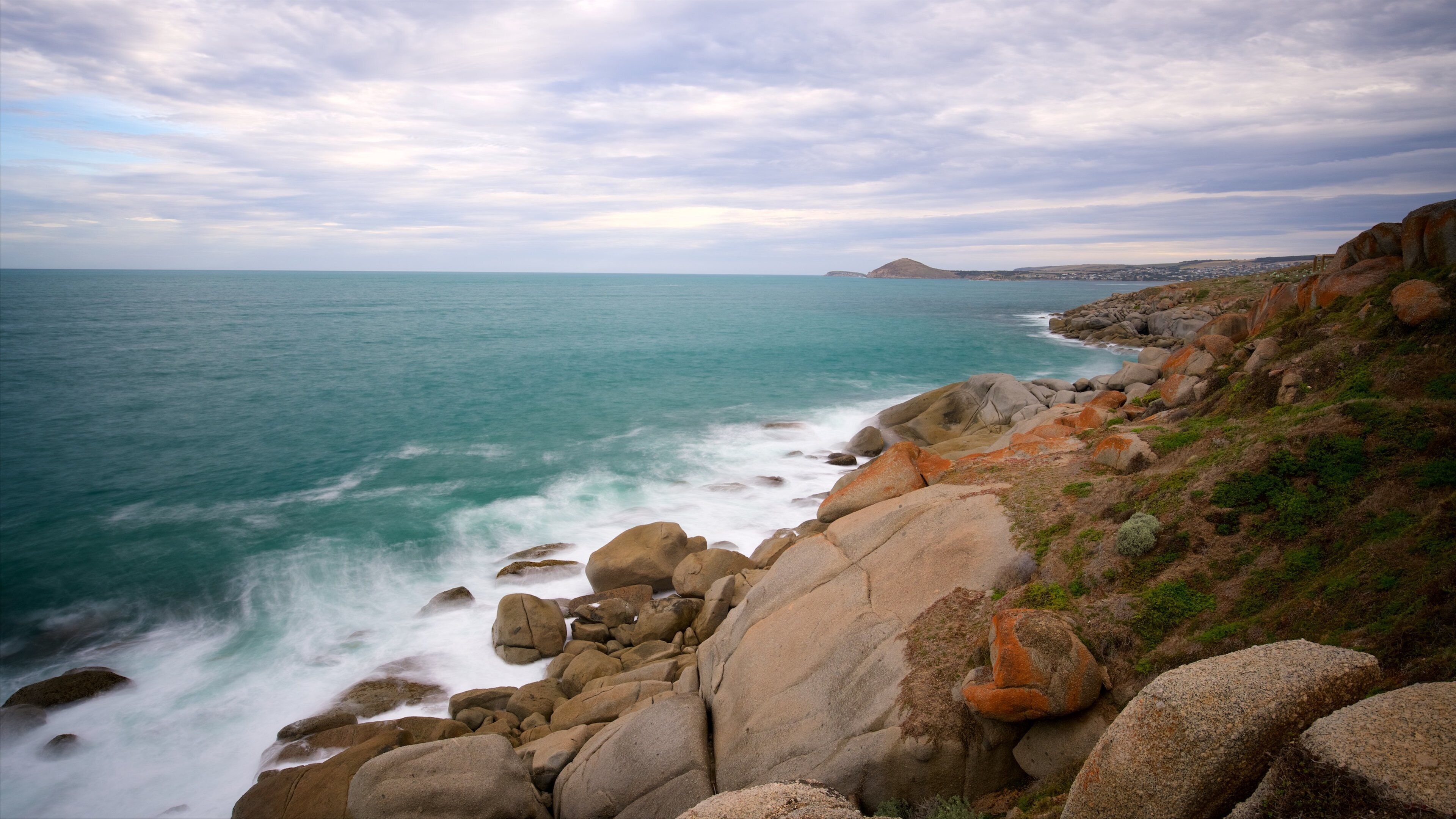 A coastal landscape with large boulders, a rocky shoreline, and a distant mountain.