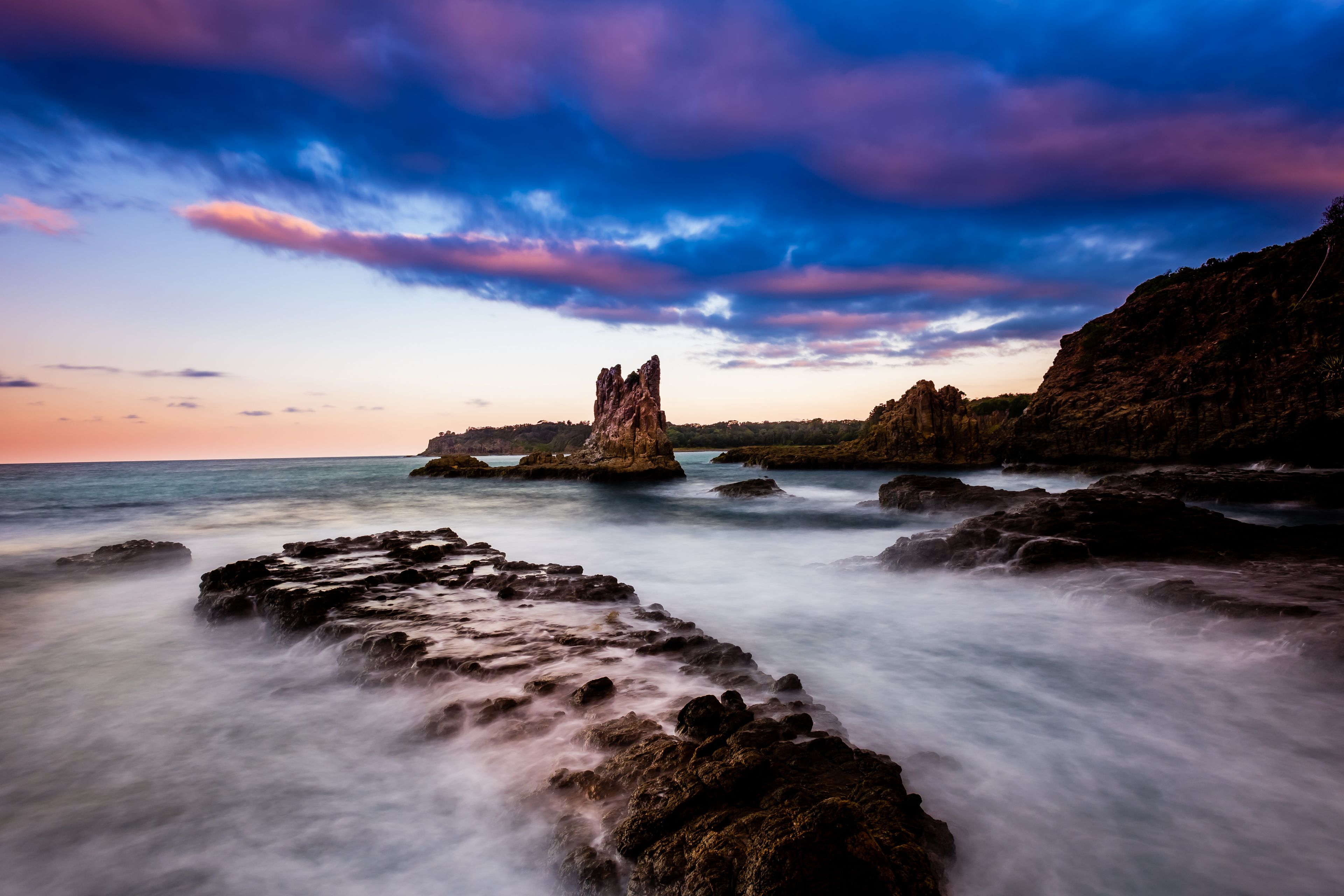 Un paysage côtier avec une formation rocheuse imposante au centre, entourée d’une mer calme et d’un ciel spectaculaire.