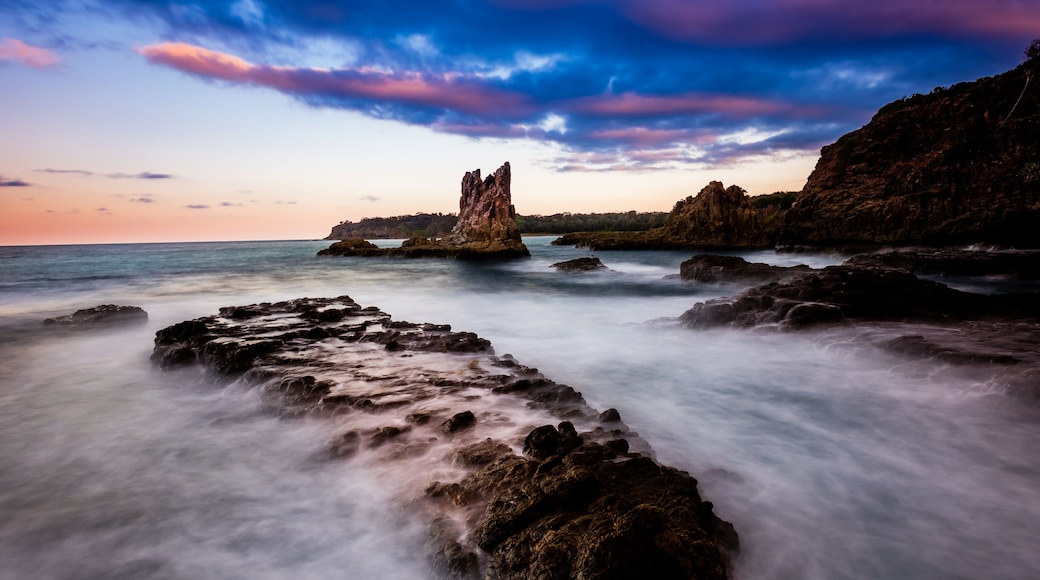 Un paysage côtier avec une formation rocheuse imposante au centre, entourée d’une mer calme et d’un ciel spectaculaire.