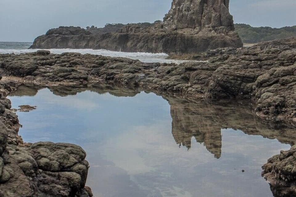 A rocky coastal landscape with a prominent rock formation in the center, surrounded by calm water reflecting the sky.
