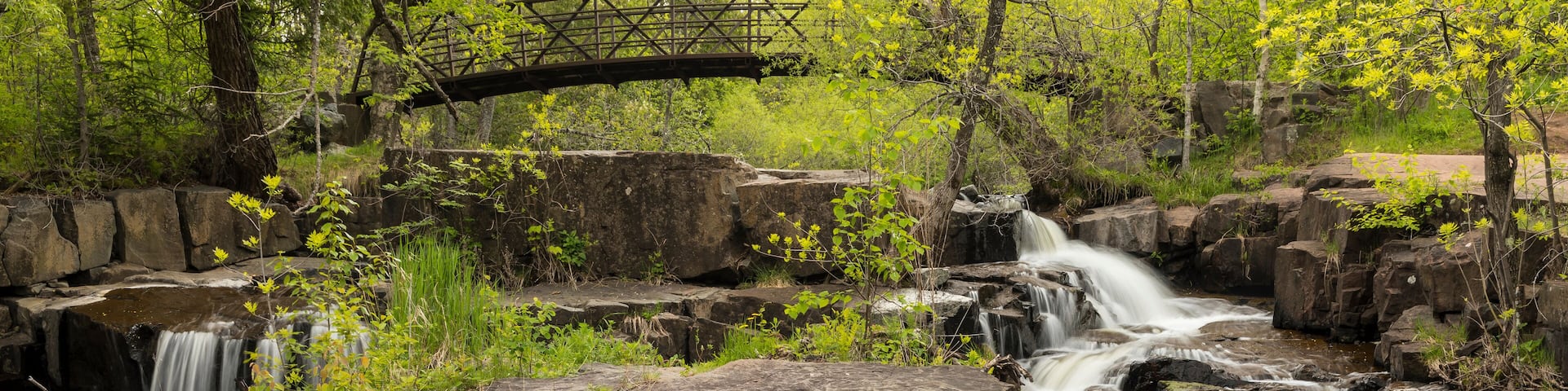 A waterfall cascading over rocks in a forested area with a bridge in the background.