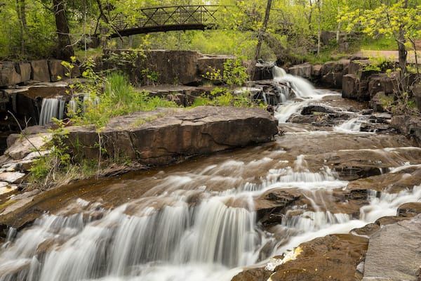 Ein Wasserfall stürzt über Felsen in einer bewaldeten Gegend, im Hintergrund ist eine Brücke zu sehen.