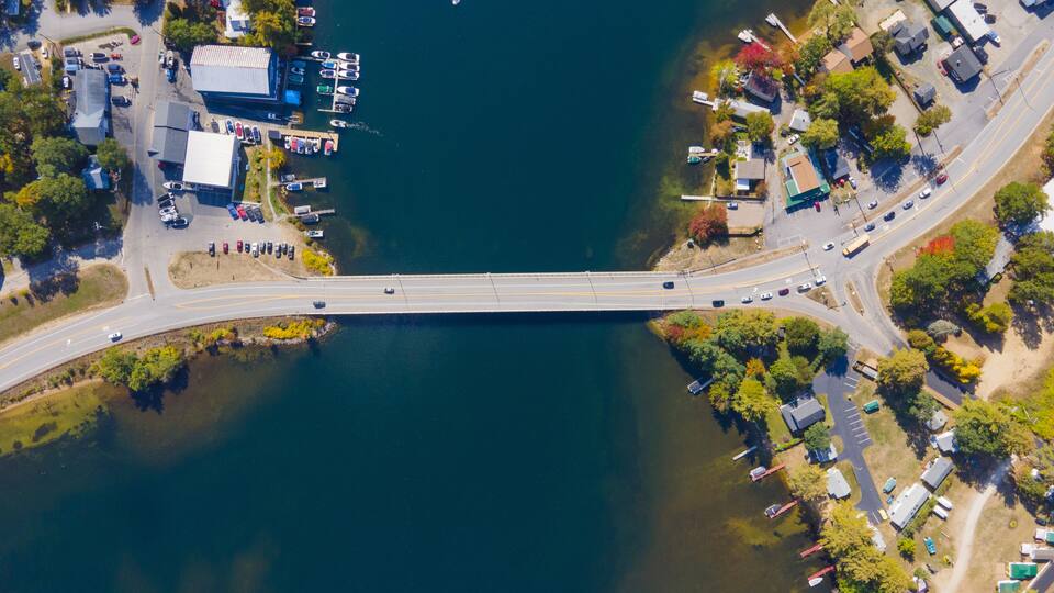 An aerial view of a bridge over a lake with boats docked on the left and a parking lot on the right.