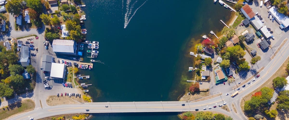 An aerial view of a bridge over a lake with boats docked on the left and a parking lot on the right.
