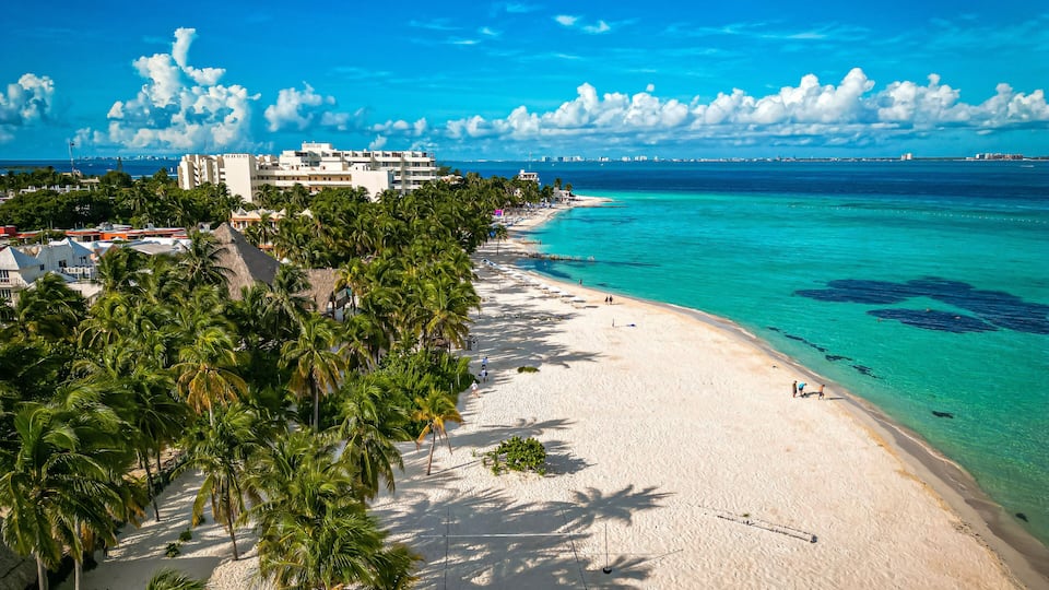 A tropical beach with clear turquoise waters, palm trees, and a resort building.