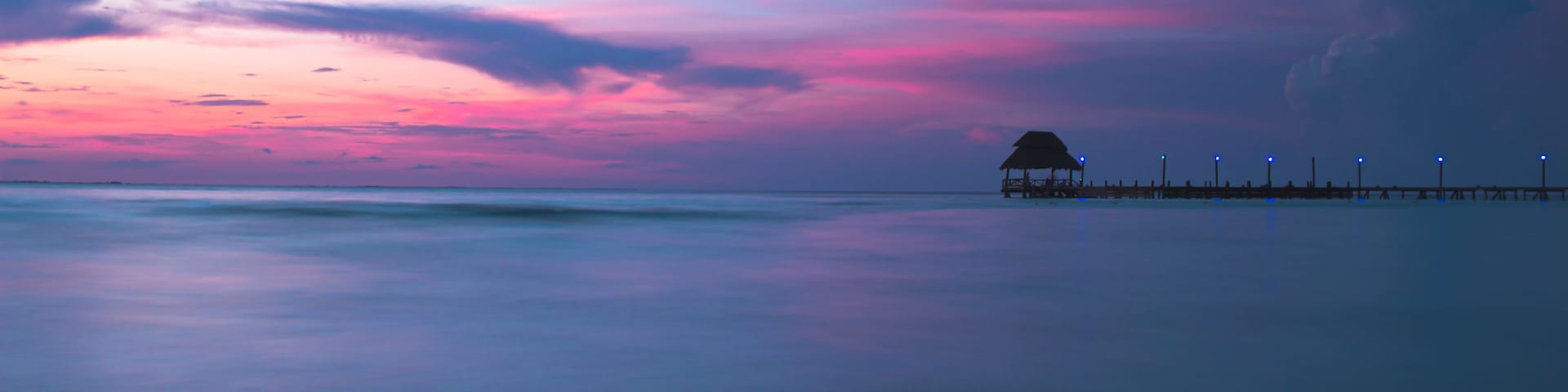 Una playa al atardecer con una cabaña de techo de paja y un muelle que se adentra en el agua.