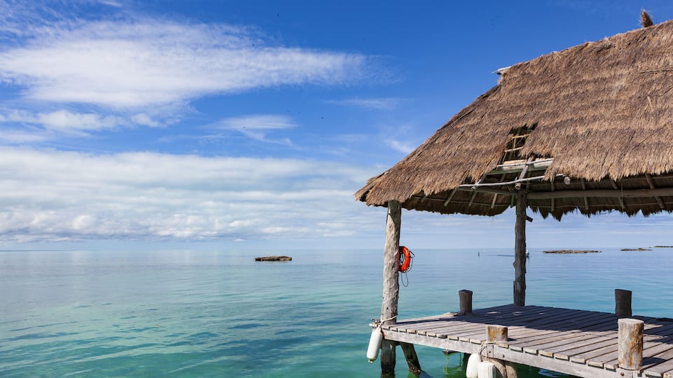 A wooden pier extending into clear turquoise water, a thatched-roof structure, and a calm sea with distant land.