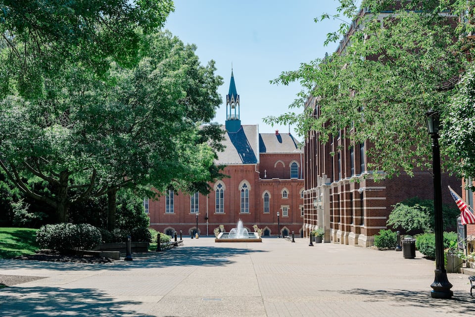 A large brick building with a blue spire, surrounded by trees and a fountain in the center.