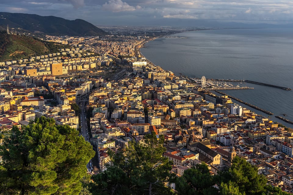 Un paisaje urbano costero con una zona densa, un muelle destacado y colinas al fondo.