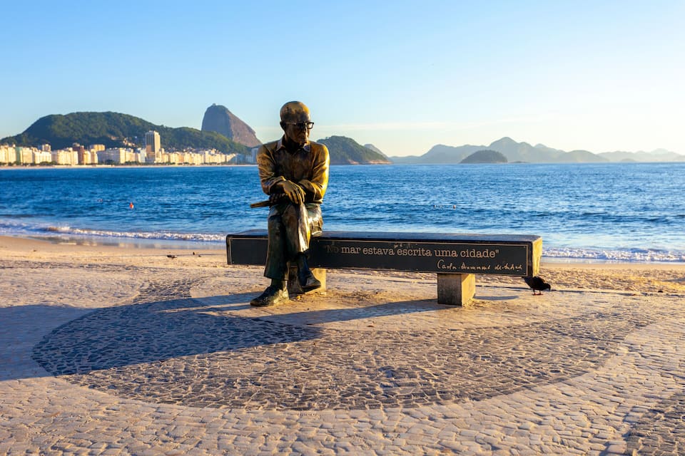 Eine Statue eines Mannes, der auf einer Bank sitzt und eine Gedenktafel trägt, mit Blick auf einen Strand, das Meer und eine Stadtlandschaft im Hintergrund.