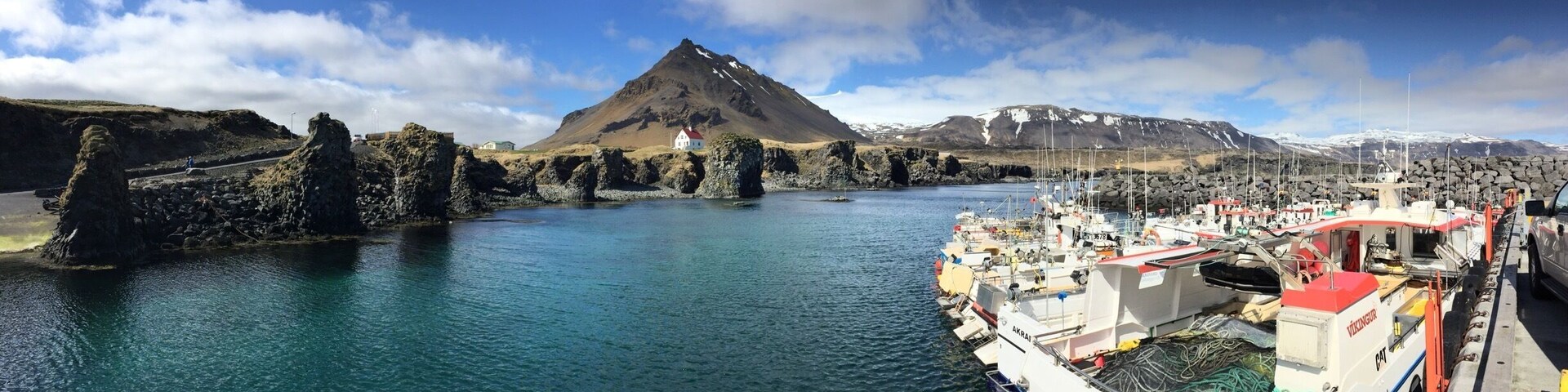Un pueblo costero con una montaña al fondo, barcos amarrados a la orilla y afloramientos rocosos en el agua.