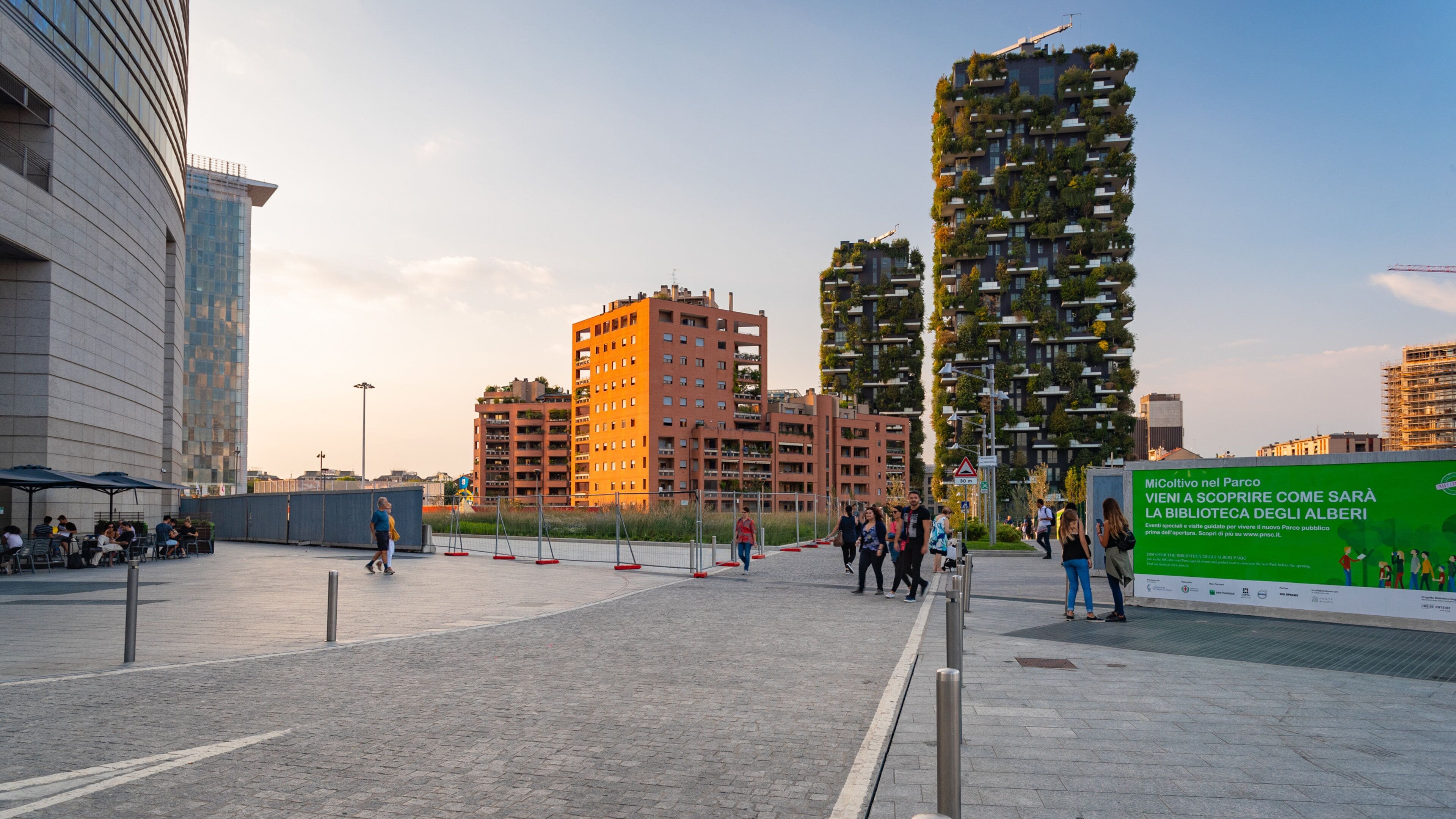 A modern urban plaza with high-rise buildings, a green advertisement board, and people walking.