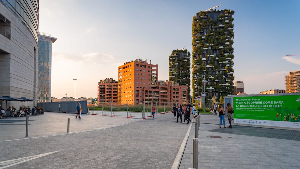 A modern urban plaza with high-rise buildings, a green advertisement board, and people walking.