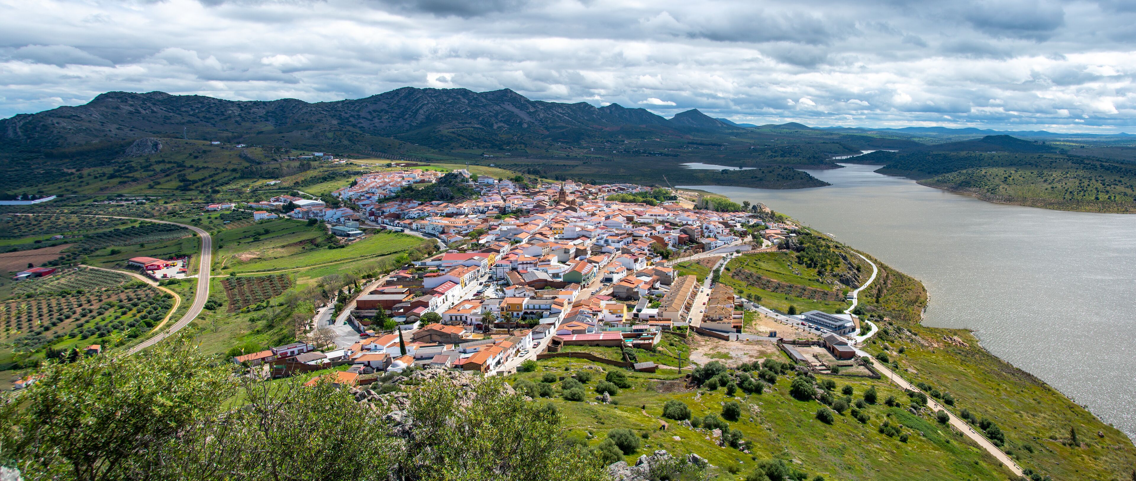 A coastal town with red-roofed buildings, a river, and mountains in the background.
