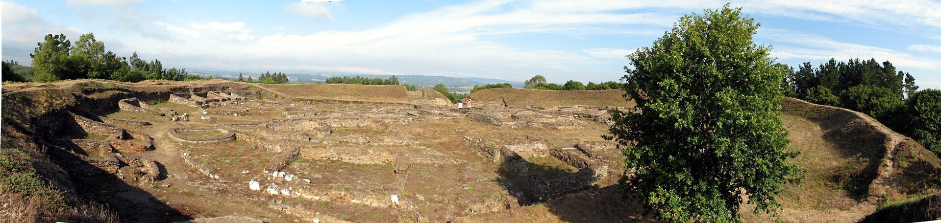 Ruinas antiguas con estructuras de piedra y senderos, rodeadas de árboles y colinas.