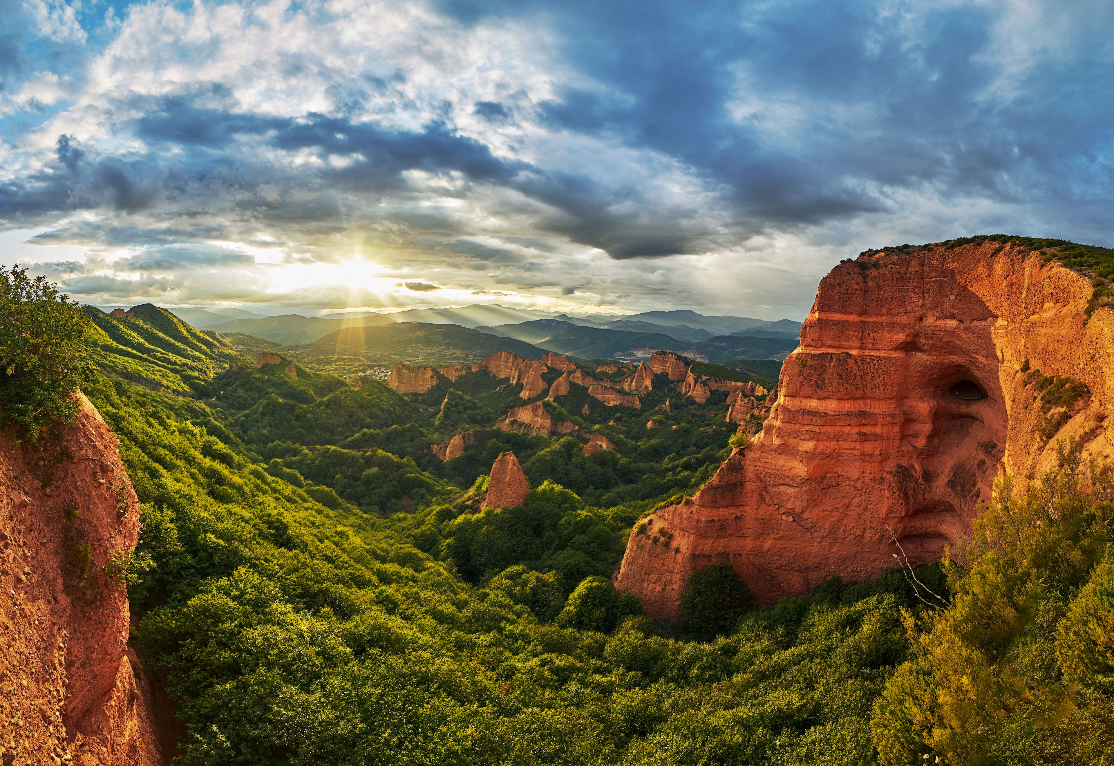 Un canyon aux formations rocheuses rouges, parsemé d’une végétation luxuriante, sous un ciel nuageux.