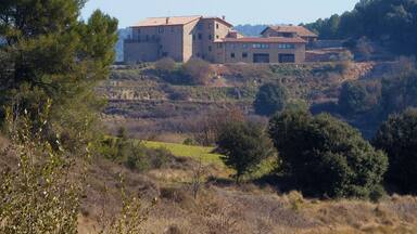 A large stone building with multiple roofs, surrounded by a hilly landscape with sparse vegetation.