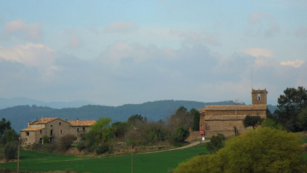Ländliche Landschaft mit einem Steingebäude und einem Turm, umgeben von grünen Feldern und sanften Hügeln in der Ferne.
