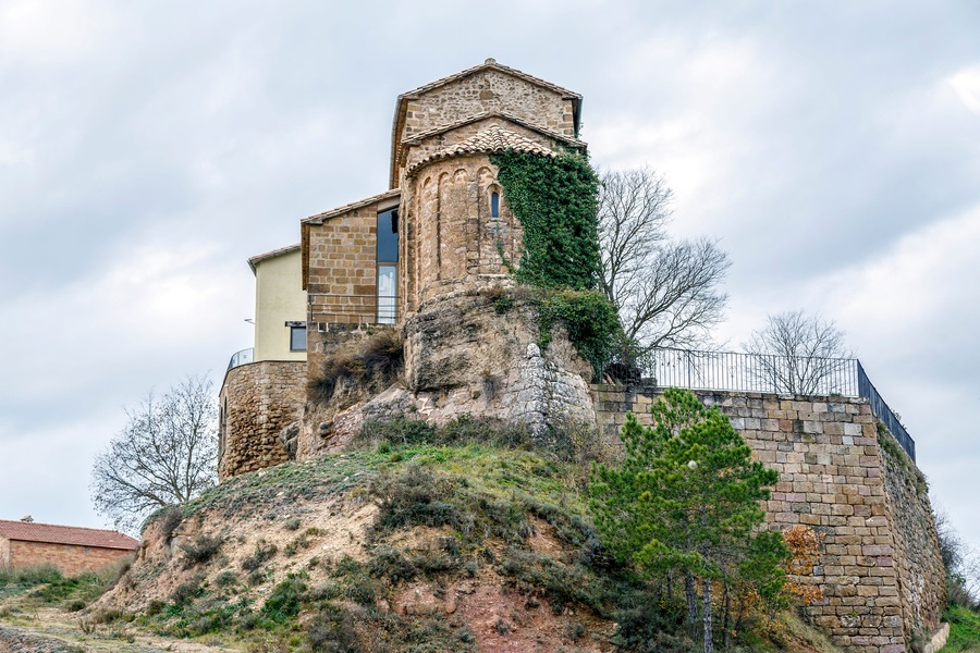 Un edificio de piedra histórico con una torre, rodeado de vegetación y un cielo nublado.