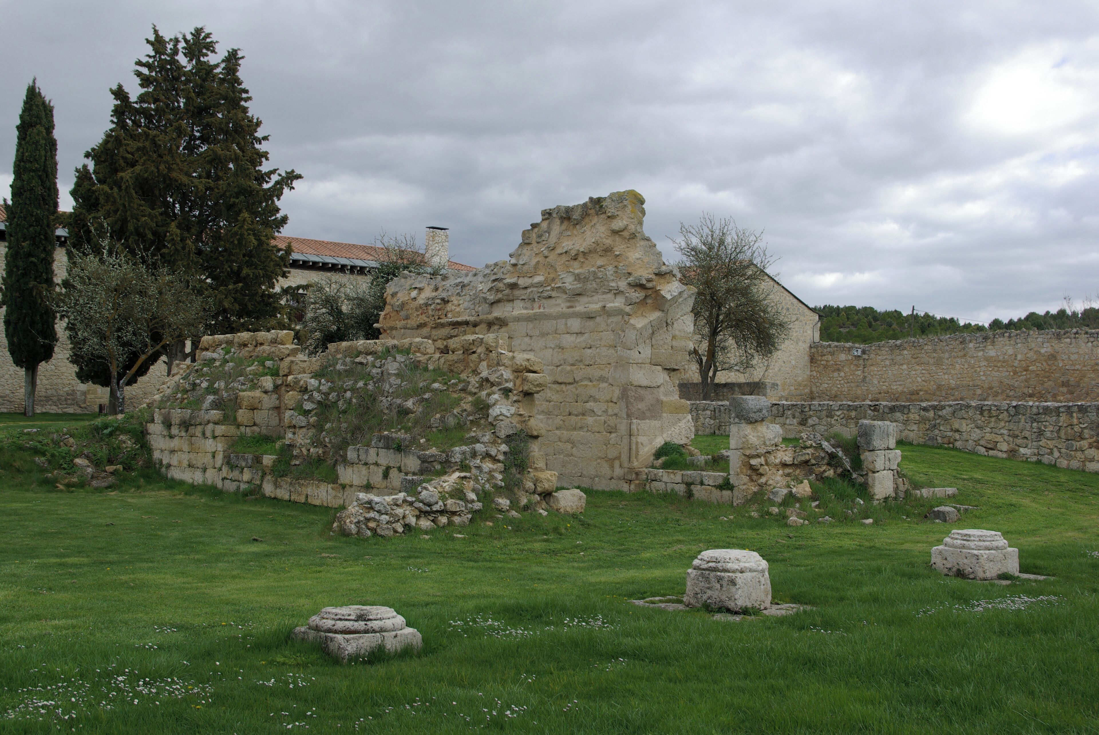 Ancient ruins with stone walls and columns, surrounded by grass and trees.