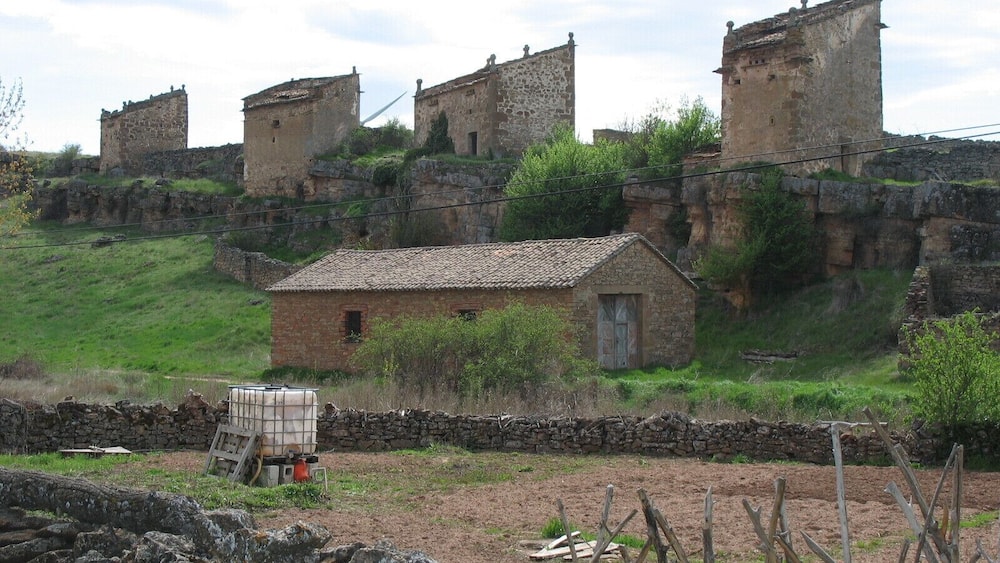 A rural landscape with a small stone house, a stone wall, and a large, ancient stone structure in the background.