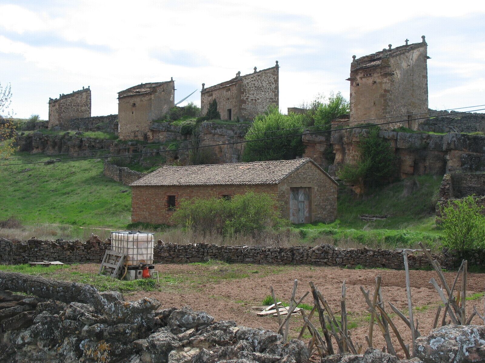 A rural landscape with a small stone house, a stone wall, and a large, ancient stone structure in the background.