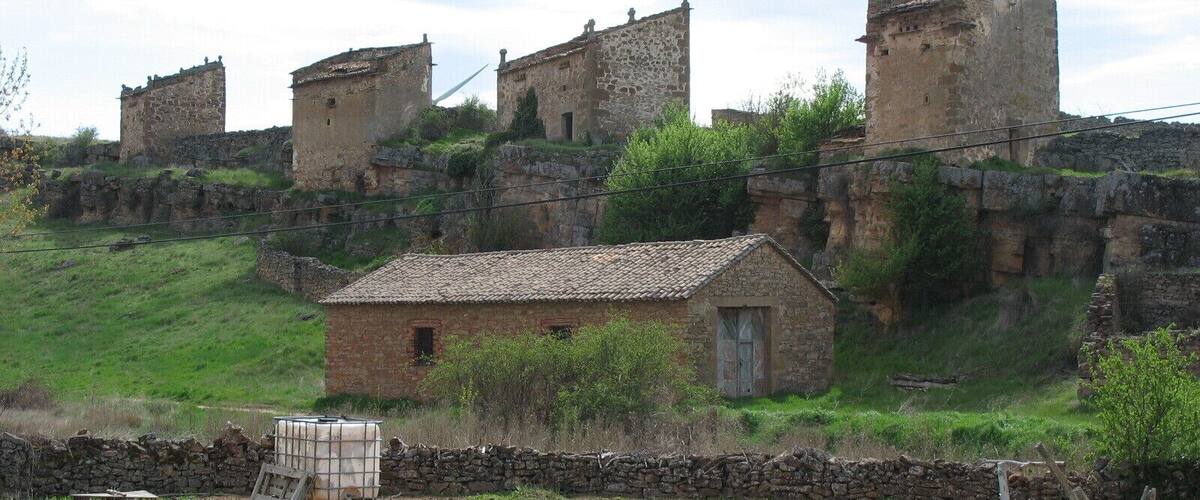 A rural landscape with a small stone house, a stone wall, and a large, ancient stone structure in the background.