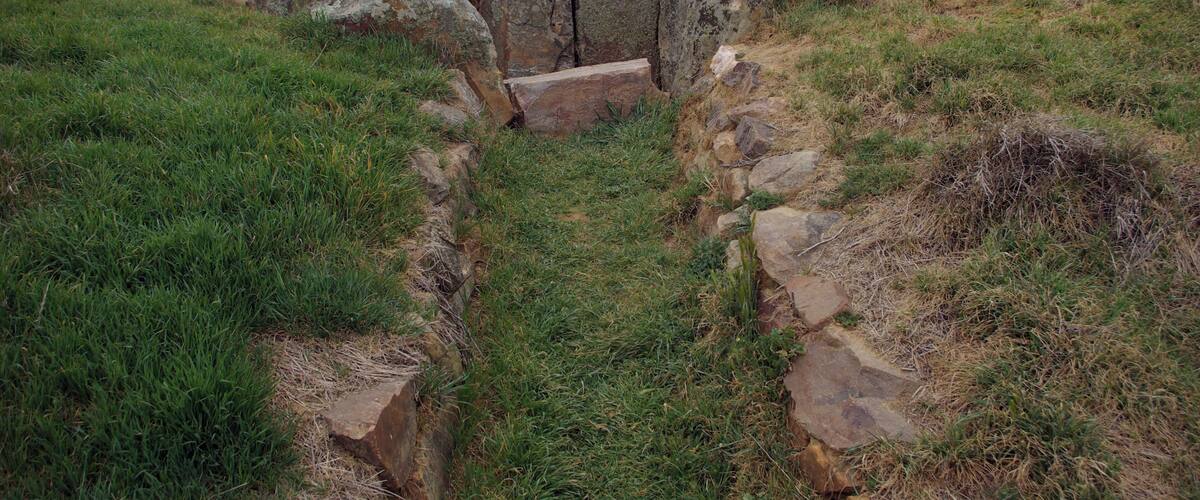 A stone structure with a narrow passageway surrounded by grass and rocks.