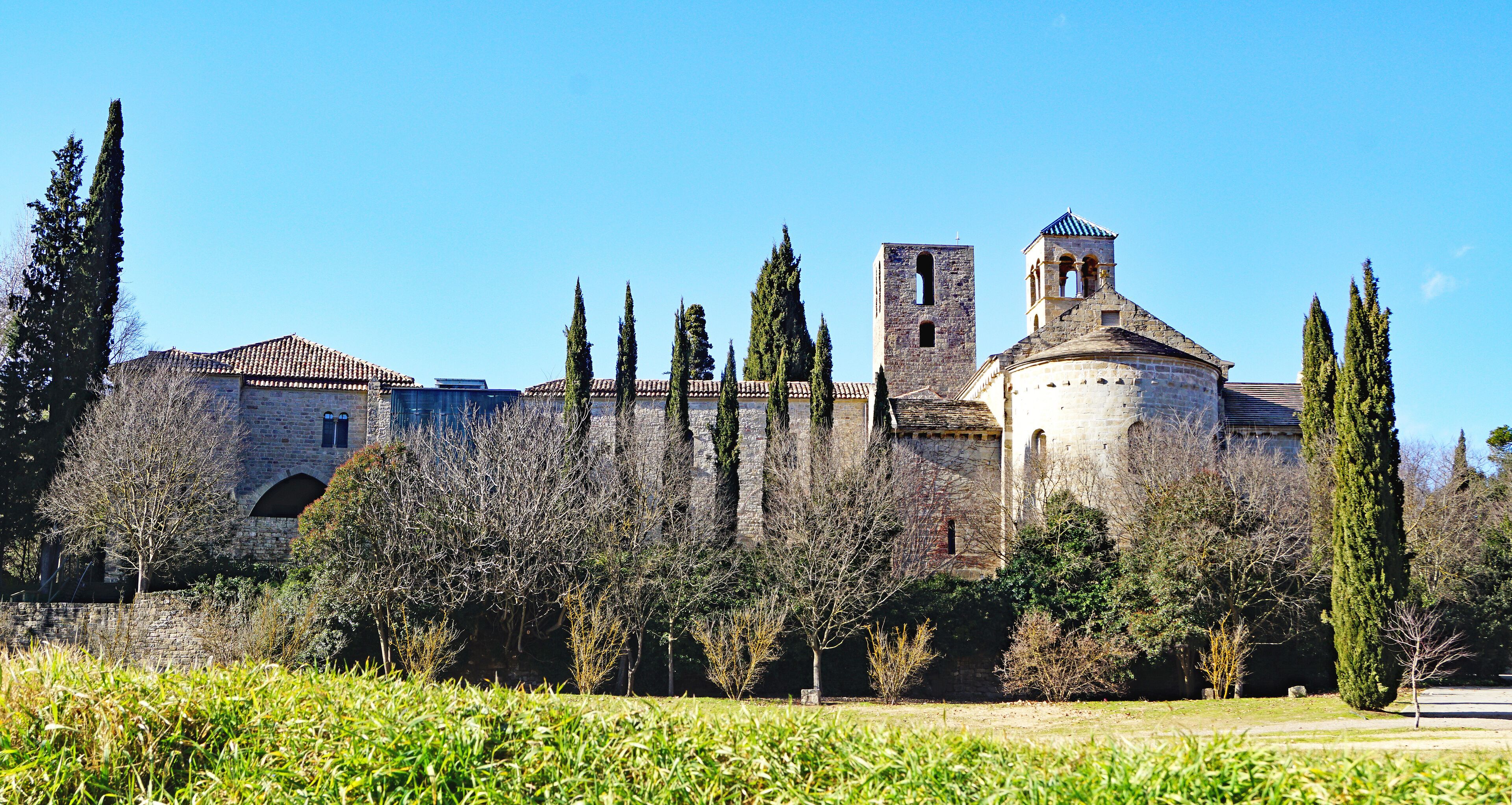 A historic stone building with a bell tower and a dome, surrounded by tall cypress trees and a grassy field.