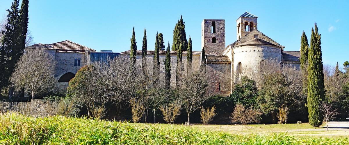 A historic stone building with a bell tower and a dome, surrounded by tall cypress trees and a grassy field.
