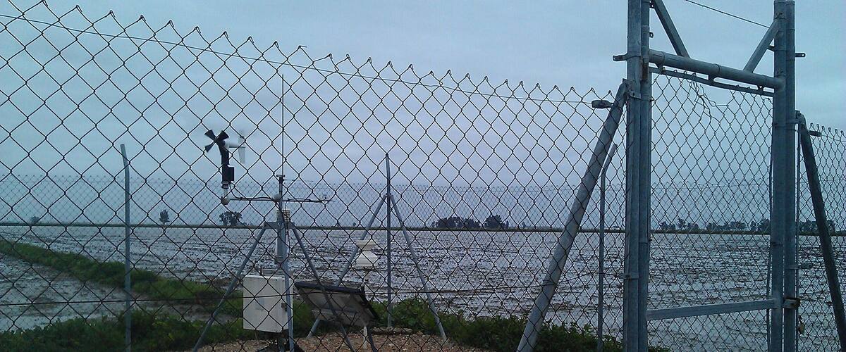 A chain-link fence with a utility pole and a bench near a body of water.