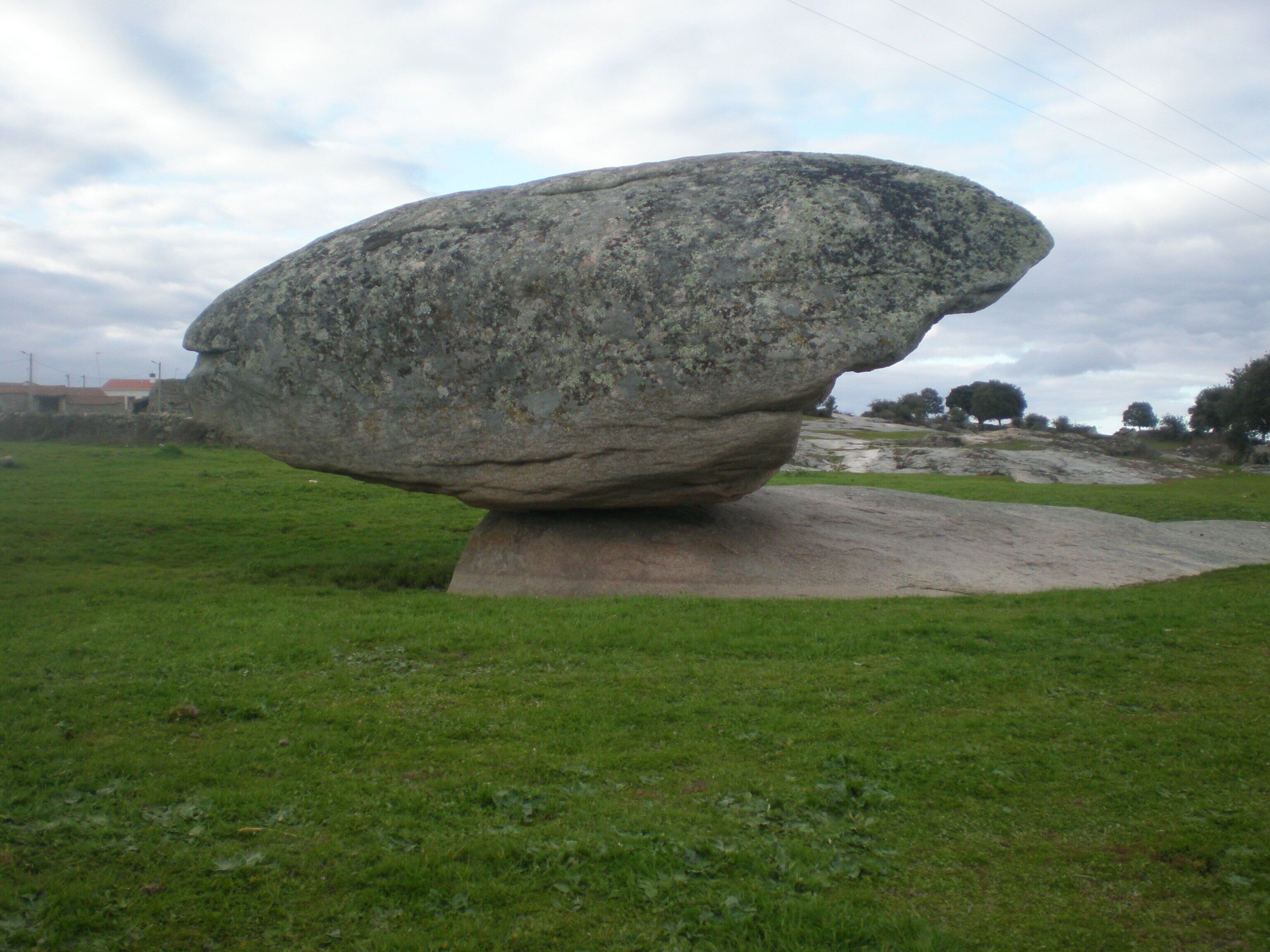 A large, balanced rock formation in a grassy field with a cloudy sky.