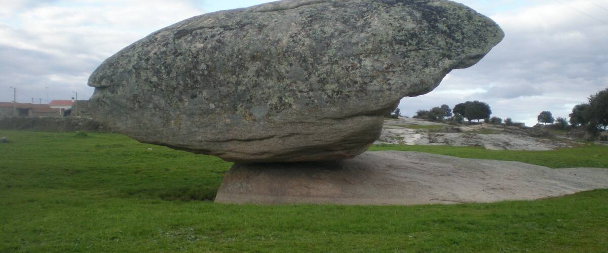 A large, balanced rock formation in a grassy field with a cloudy sky.