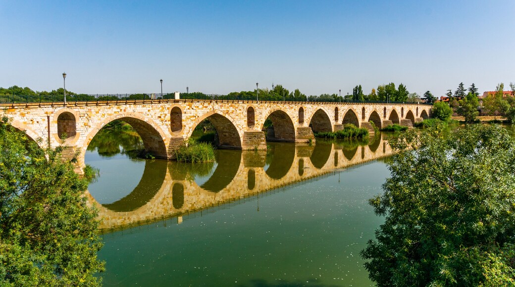 Un puente de piedra con varios arcos que cruza un río tranquilo, rodeado de abundante vegetación.