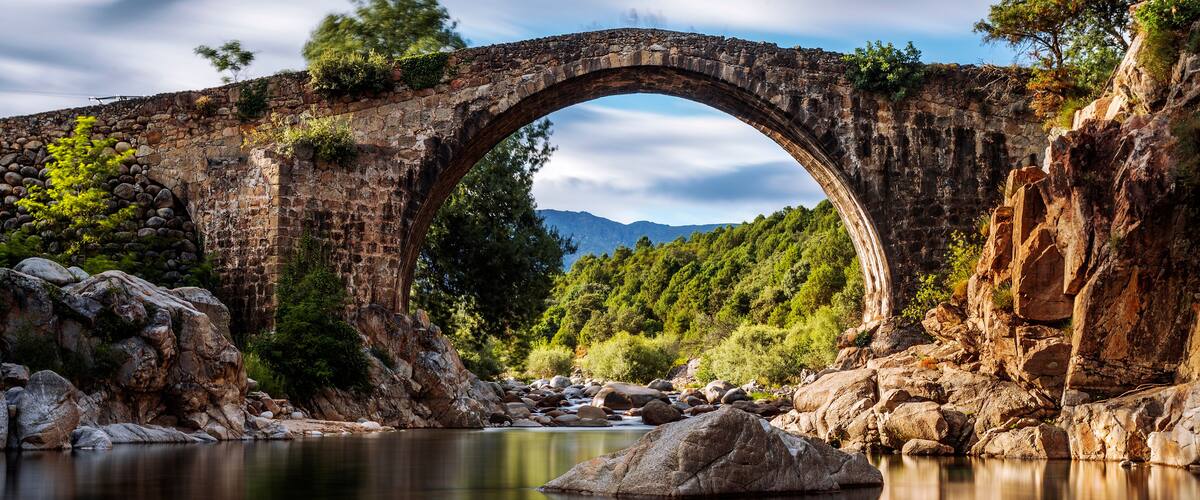 A stone bridge over a river, surrounded by rocky terrain and trees.