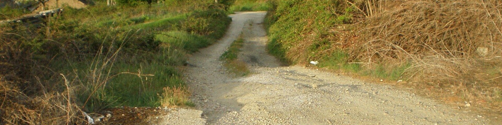 Un camino de grava serpentea por un paisaje rural con poca vegetación y algunas casas dispersas.