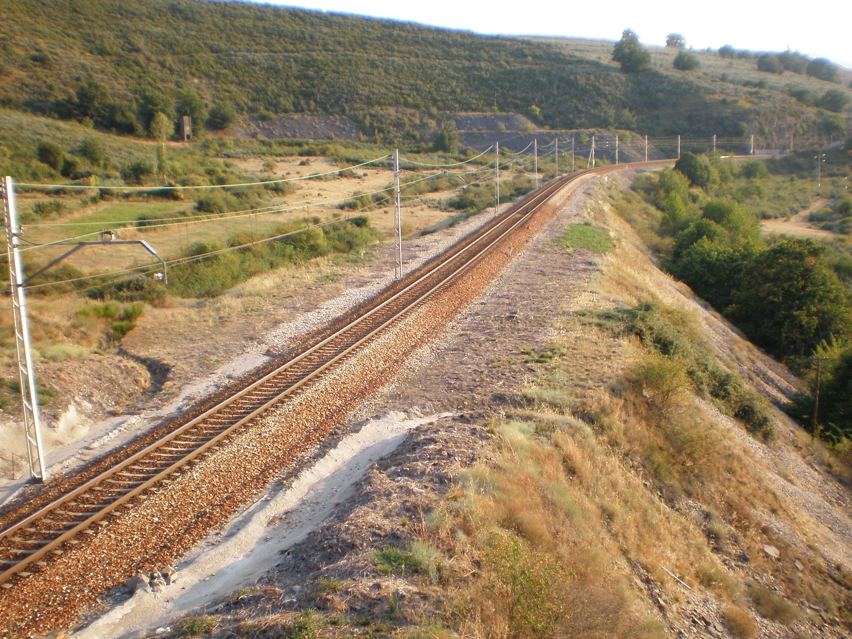 A railway track curving through a hilly landscape with sparse vegetation and a clear sky.