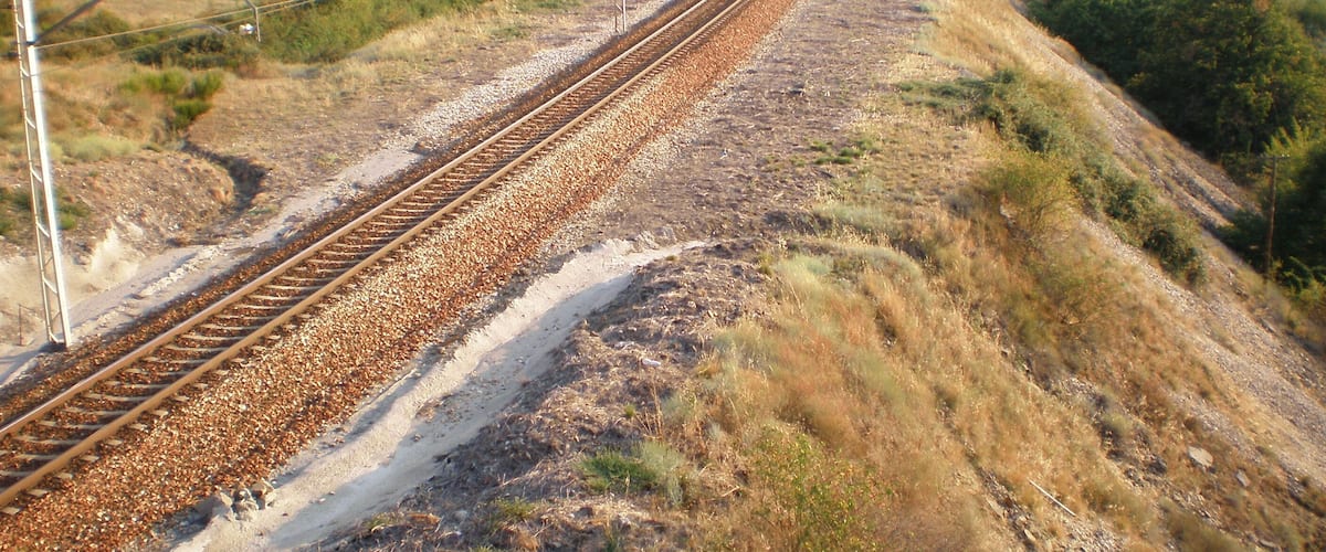 A railway track curving through a hilly landscape with sparse vegetation and a clear sky.