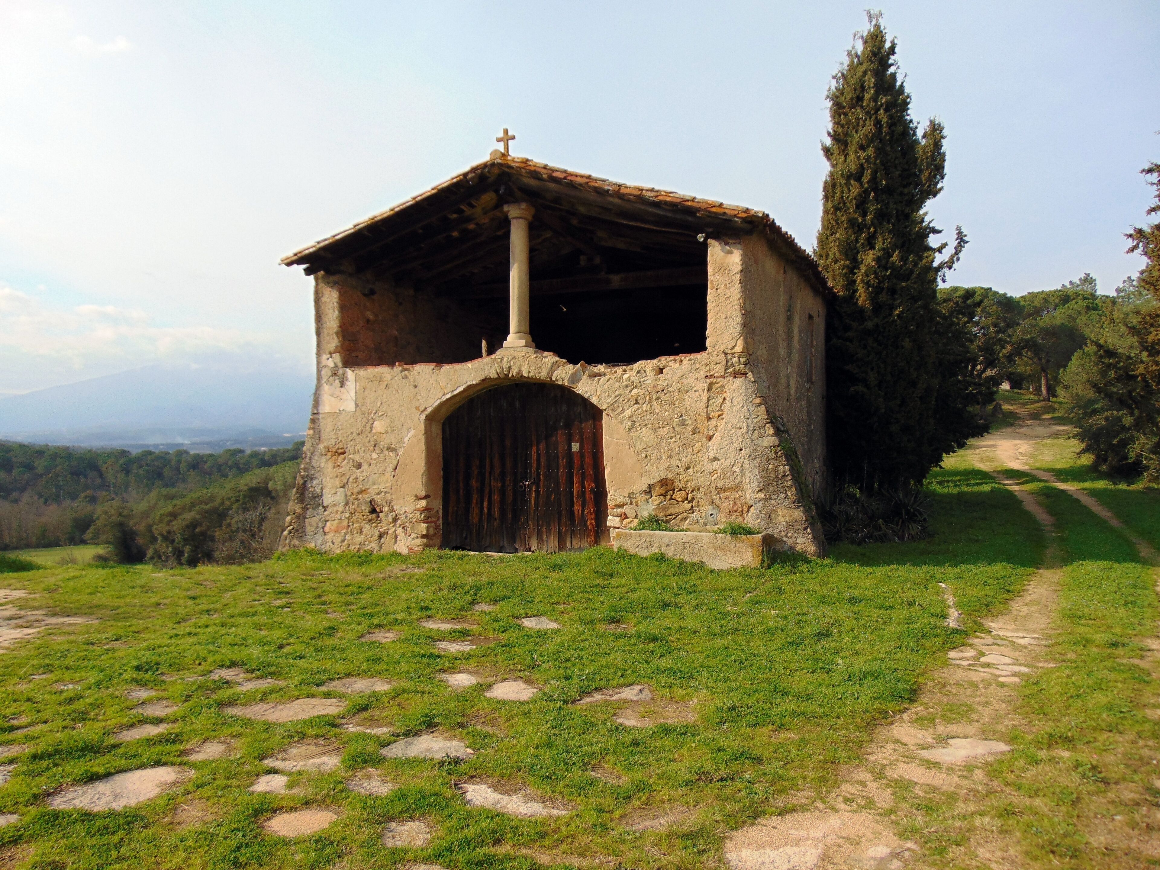 A small stone chapel with a cross on top, surrounded by a grassy area with a dirt path leading to it.