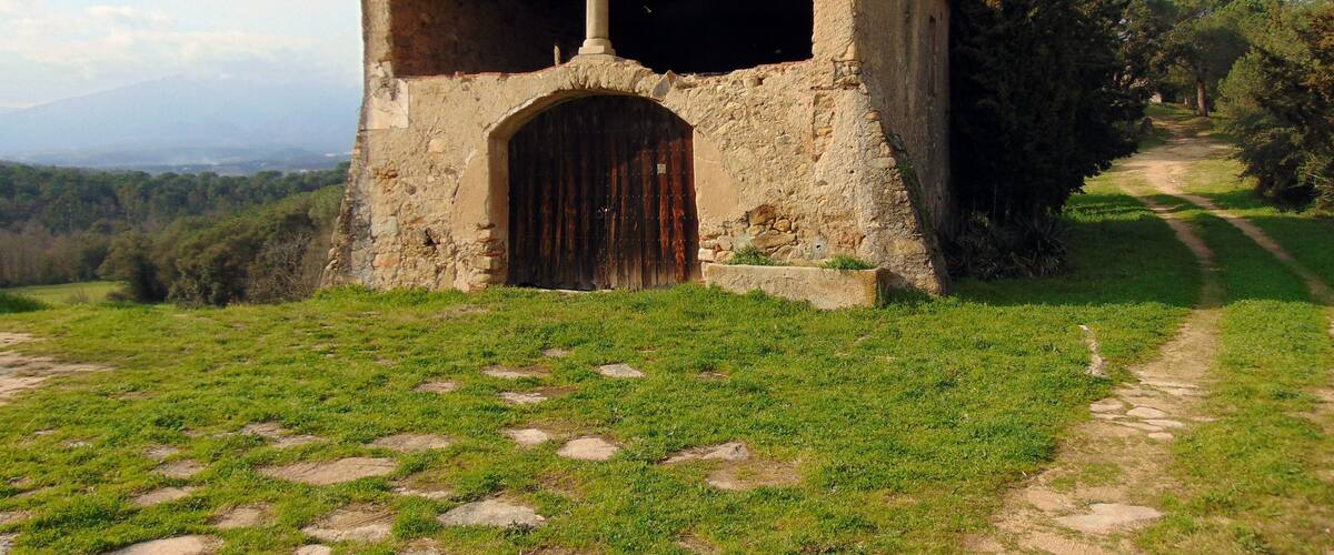A small stone chapel with a cross on top, surrounded by a grassy area with a dirt path leading to it.