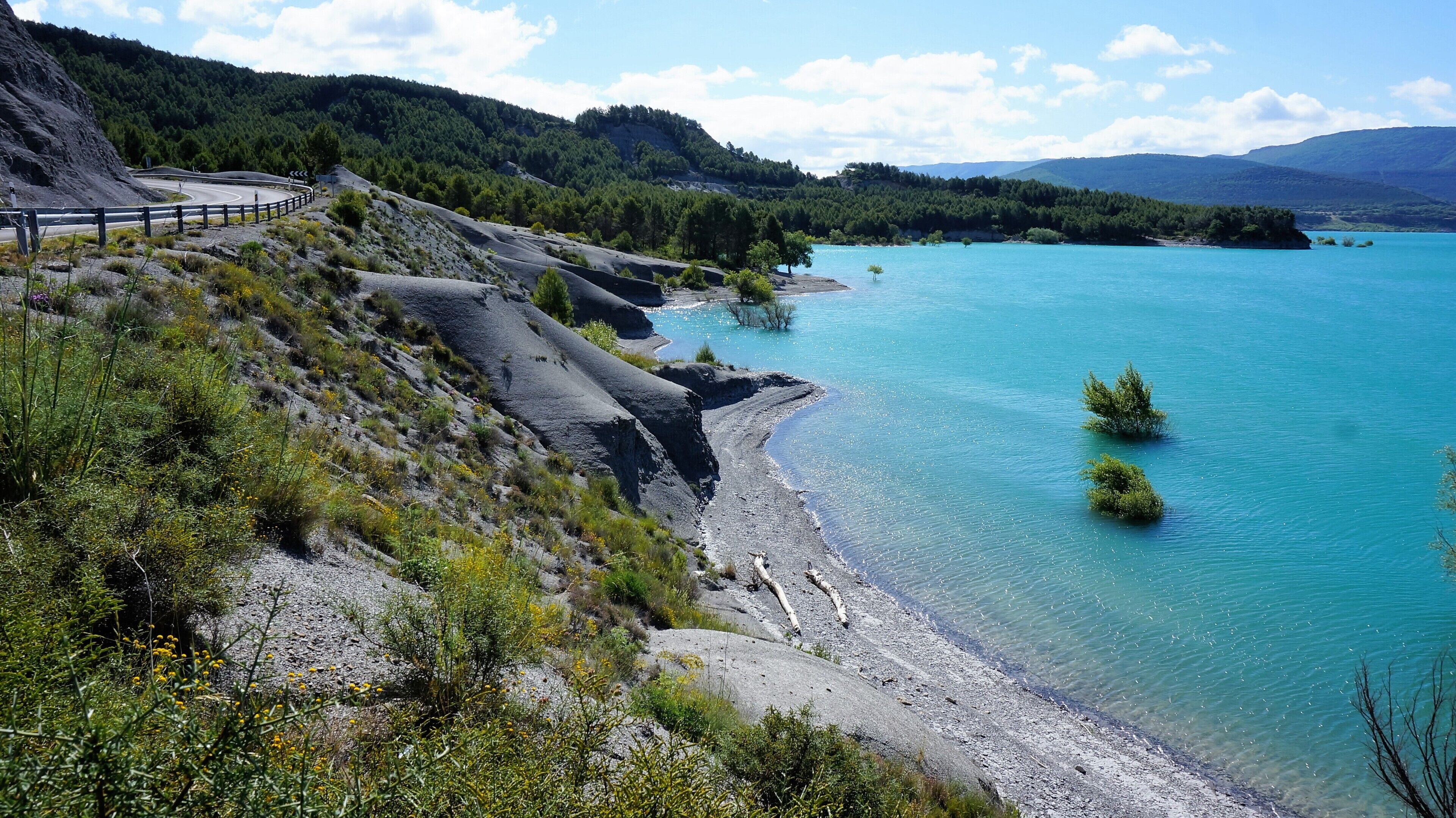 A winding road along a coastal area with clear turquoise waters, surrounded by green hills and sparse vegetation.