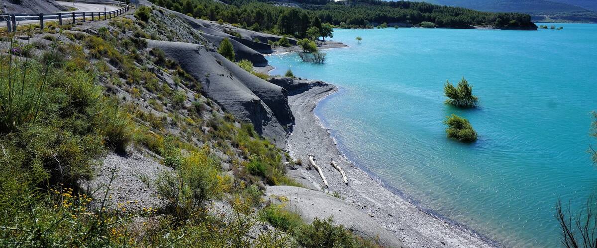 A winding road along a coastal area with clear turquoise waters, surrounded by green hills and sparse vegetation.