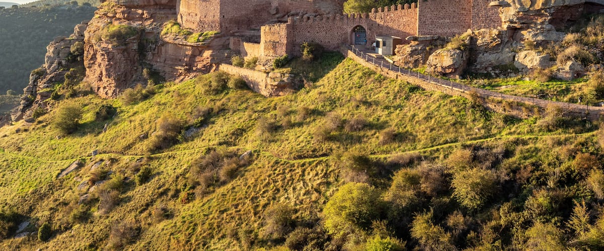 A historic stone fortress on a hill with surrounding greenery and a clear sky.