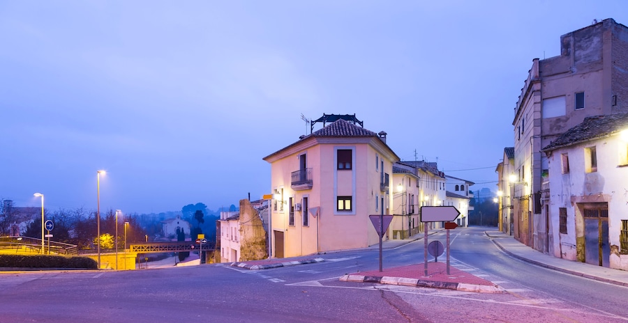 A quiet street at dusk with historic buildings, a pedestrian bridge, and street lamps.