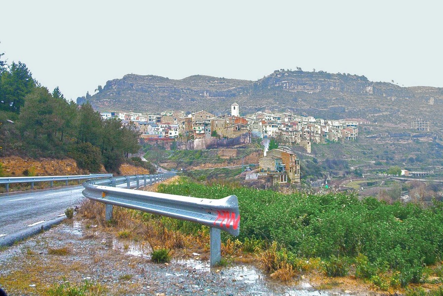Una carretera costera con barandilla, un pueblo con edificios de colores y una colina al fondo.